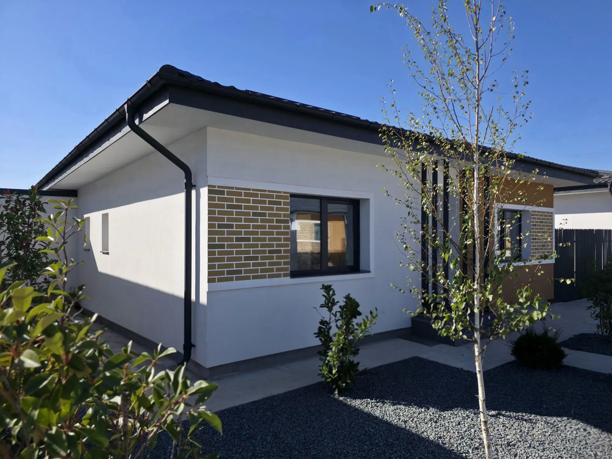 Casa Parter finalizată în Tunari — colțul casei, cu fereastra placată cu cărămidă, mesteacănul și grădina cu pietriș